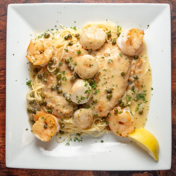Italian pasta dish with chicken, shrimp, scallops, capers, and lemon wedge in a creamy sauce.